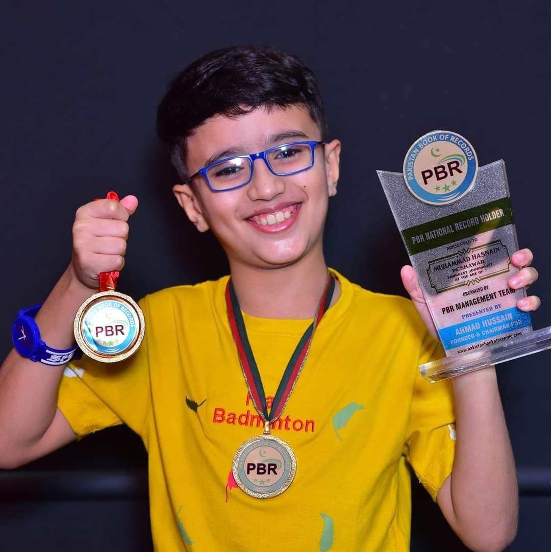 Smiling child with glasses holding a trophy and medal against a dark background.